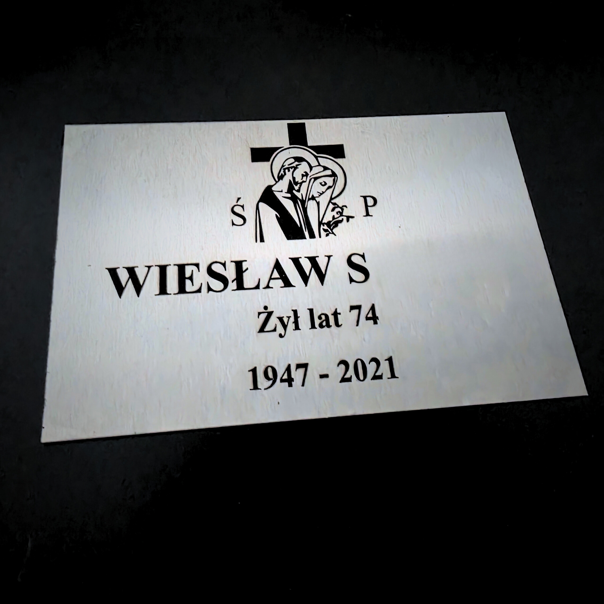 Zdjęcie przedstawia grawerowaną tabliczkę nagrobną. Na tabliczce można zobaczyć Imię, pierwszą literę nazwiska, wiek oraz rok urodzenia i śmierci.