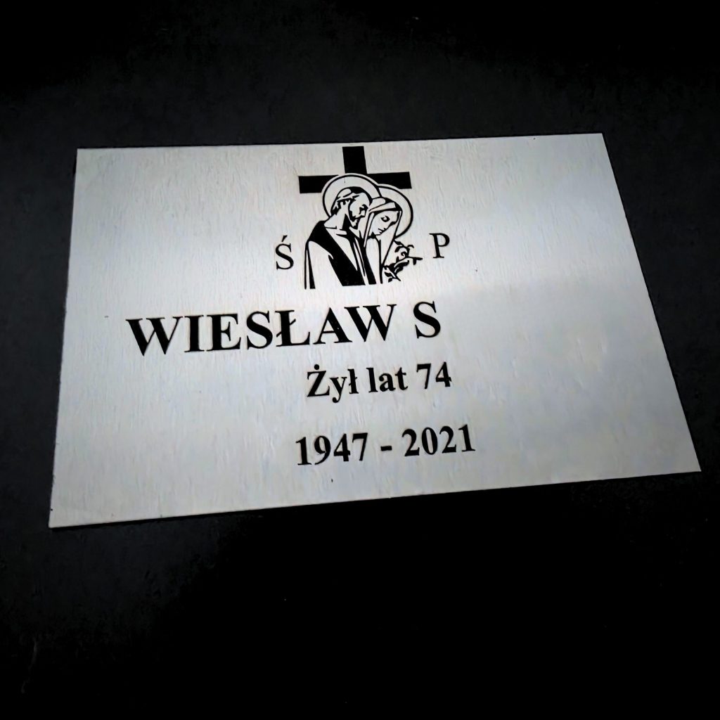 Zdjęcie przedstawia grawerowaną tabliczkę nagrobną. Na tabliczce można zobaczyć Imię, pierwszą literę nazwiska, wiek oraz rok urodzenia i śmierci.