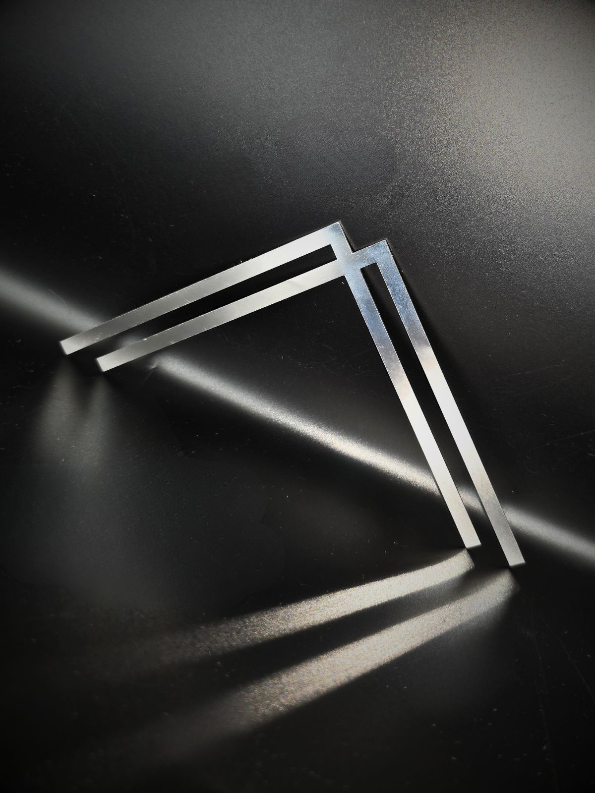 Zdjęcie przedstawia ozdoby ornament montowany w rogu płyty pomnika dla zamknięcia kompozycji. Ornament przedstawia przecinające się w jednym punkcie linie w kształcie "L". Stoi oparte o czarne tło i rzuca czarny długi cień na podstawę.