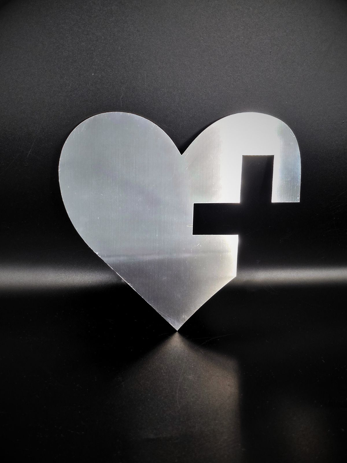 Zdjęcie przedstawia serce wycięte ze stali nierdzewnej. W sercu widać fragment krzyża. Stoi oparte o czarne tło i rzuca cień przed siebie.