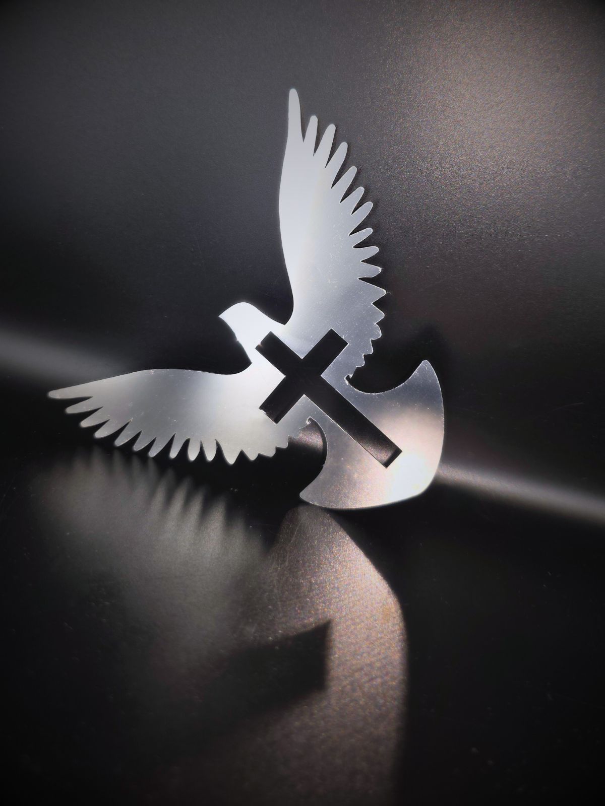 Zdjęcie przedstawia gołębia z krzyżem w ciele. Gołąb ma rozpostarte skrzydła. Ozdoba wykonana jest ze stali nierdzewnej, postawiona na czarnym tle rzuca cień na czarną podłogę.