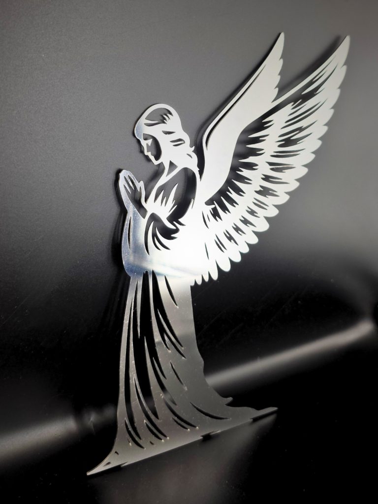 Zdjęcie przedstawia figurę anioła stojącego bokiem ze złożonymi rękami. Detal wykonany ze stali nierdzewnej stoi oparty o czarne tło.