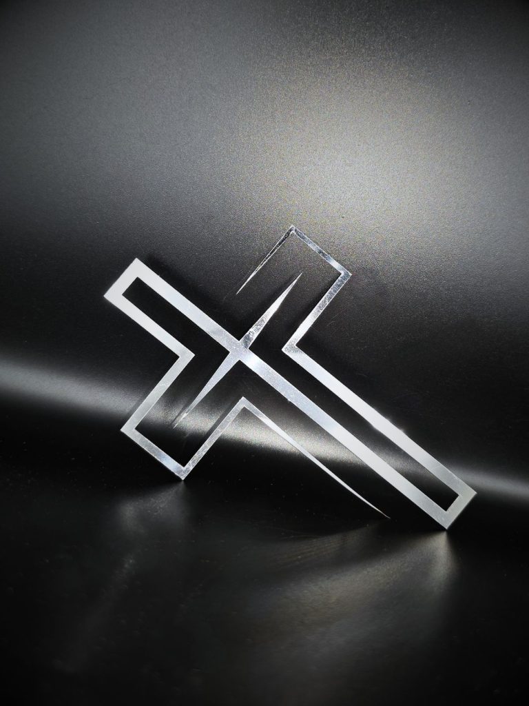 Zdjęcie przedstawia leżący na boku bardzo nowocześnie wyglądający krzyż z cienką nóżką. Krzyż opiera się o jedno ramię i czarne tło. Rzuca na ziemie cień.