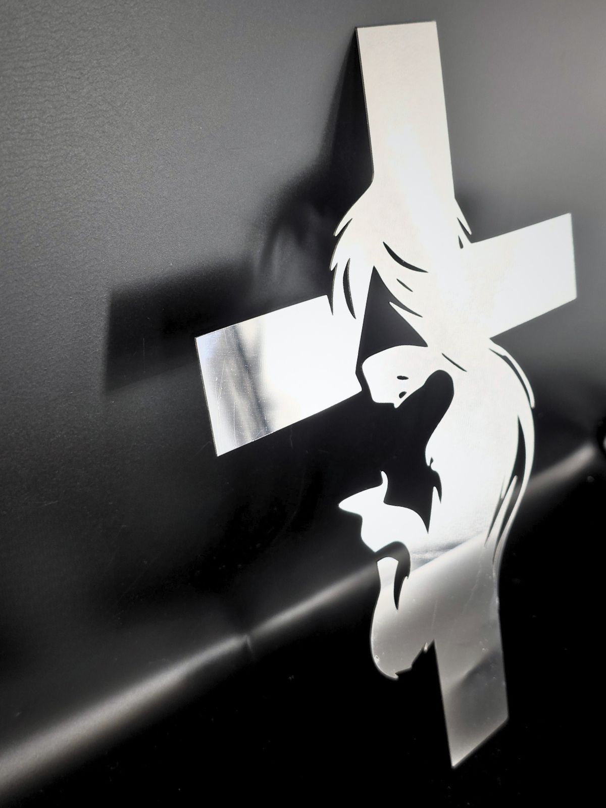 Zdjęcie przedstawia twarz Jezusa zaprojektowaną w krzyż. Krzyż stoi oparty o czarną ścianę, rzuca na nią cień. Krzyż wykonany jest ze stali nierdzewnej, w której odbijają się fragemnty otoczenia.