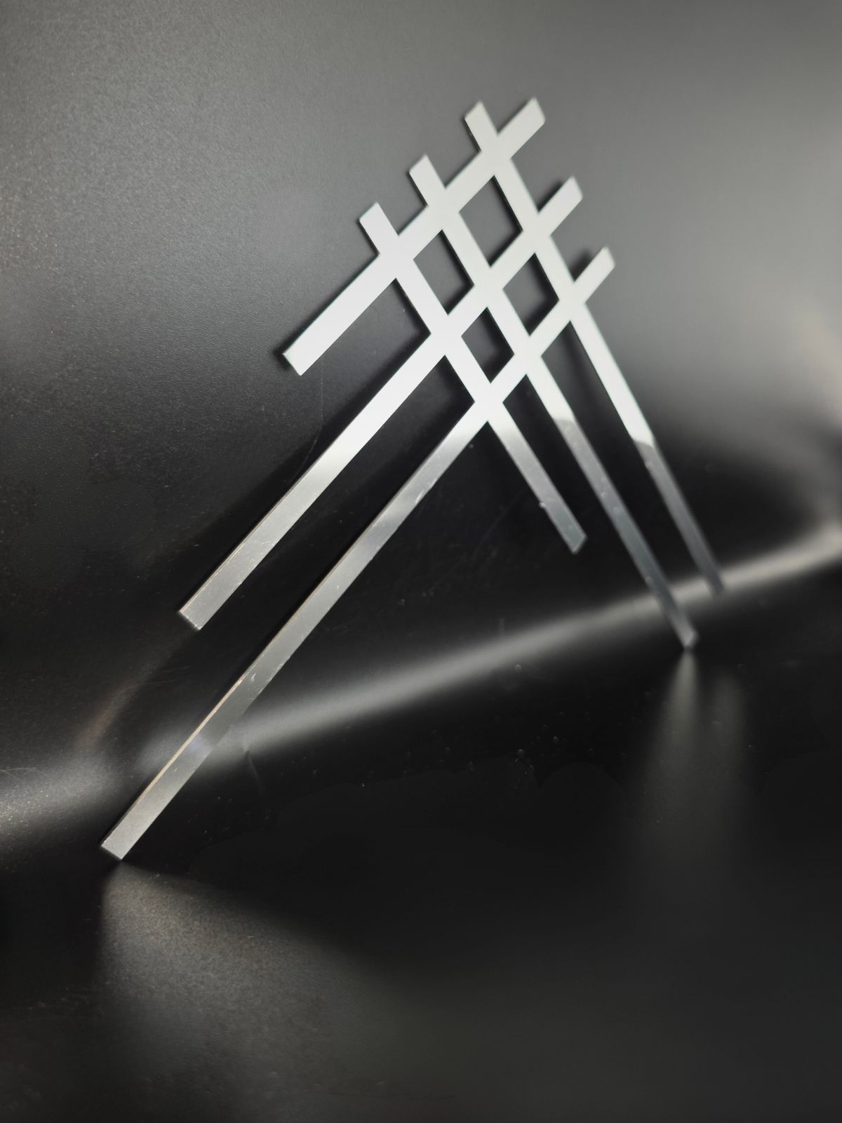 Zdjęcie przedstawia ozdobną listwę na pomnik wykonaną ze stalii nierdzewnej. Listwa składa się z 6 różnych i przecinających się lini, które w ten sposób w miejscu przecięcia tworzą 4 kwadraty.